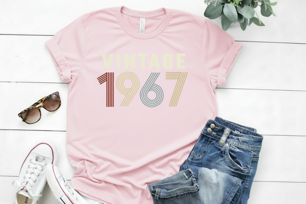Pink t-shirt with 'Vintage 1967' text, sunglasses, jeans, and sneakers on a white wooden surface.