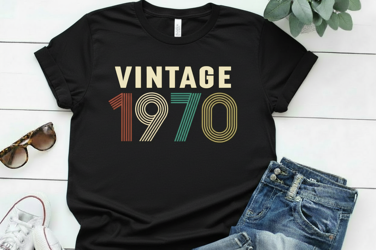 Black t-shirt with 'Vintage 1970' text on a white wooden surface with sunglasses and jeans.
