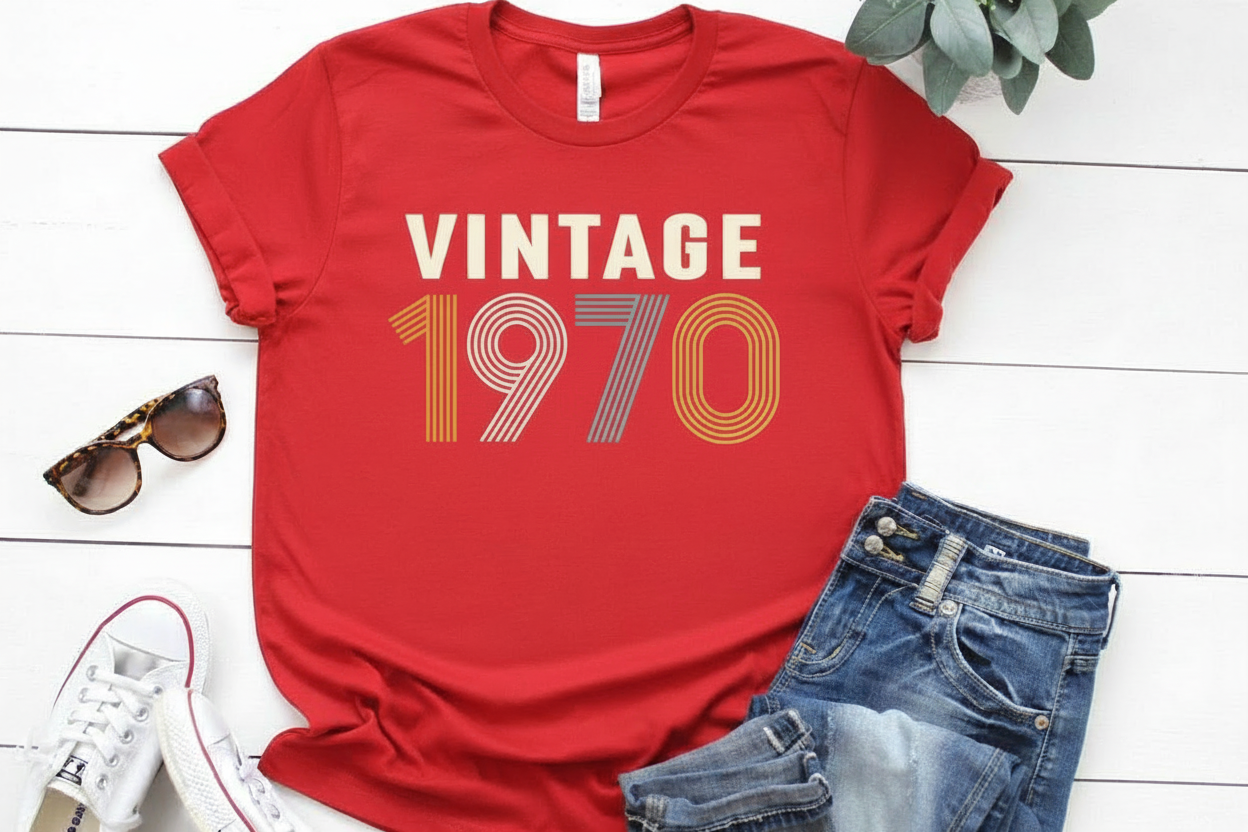 Red t-shirt with 'Vintage 1970' text, sunglasses, and jeans on a white wooden surface.