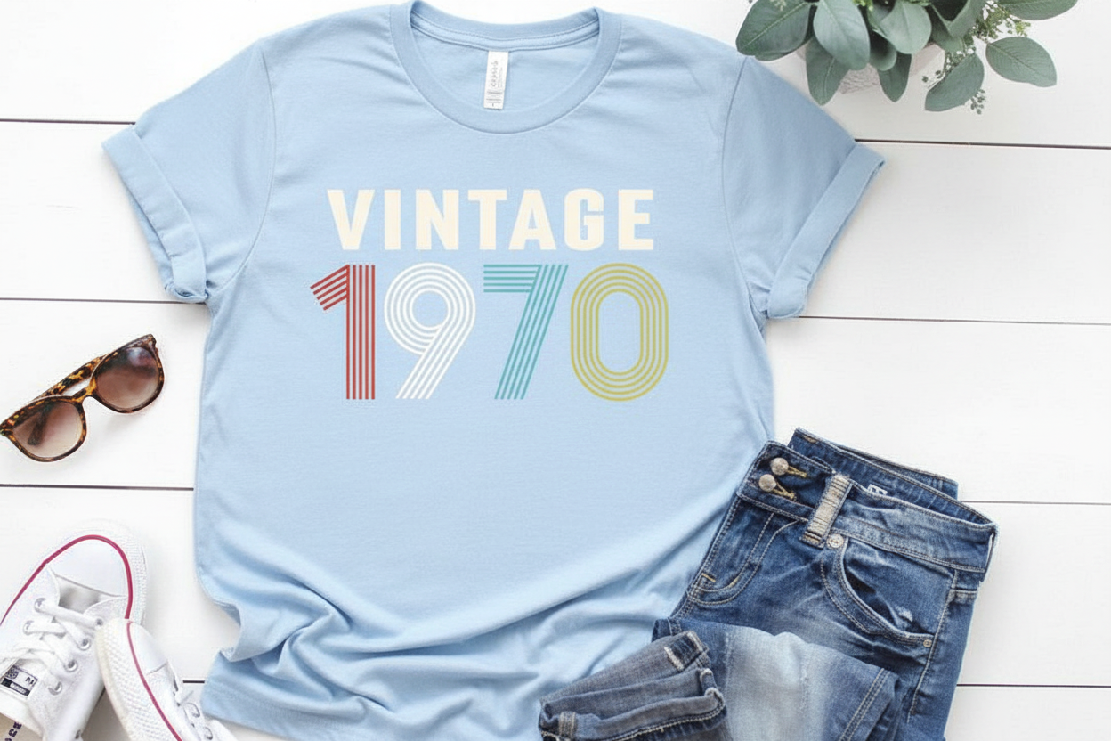 Light blue t-shirt with 'Vintage 1970' text, sunglasses, jeans, and sneakers on a white wooden surface.