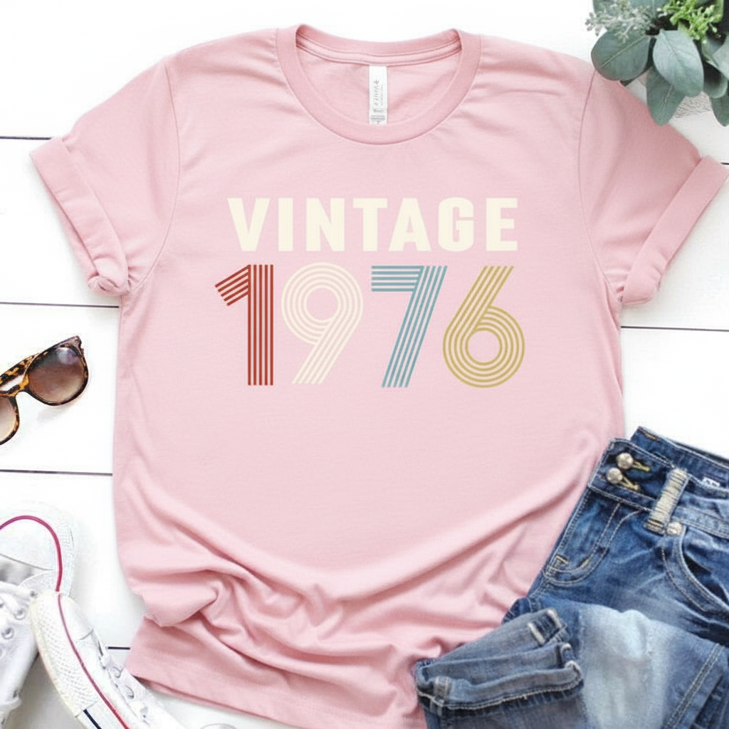 Pink t-shirt with 'Vintage 1976' text on a white surface with sunglasses and jeans.