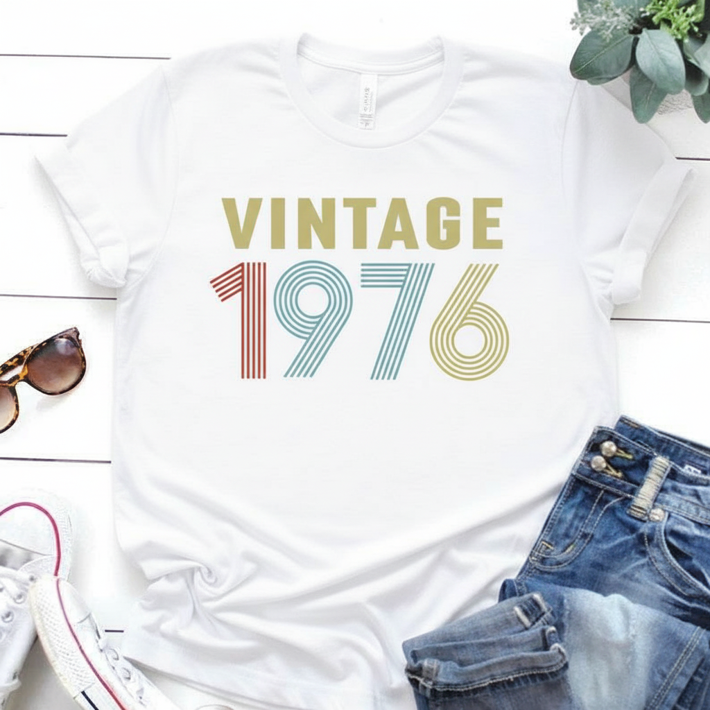 White t-shirt with 'Vintage 1976' design on a wooden surface with sunglasses and jeans.