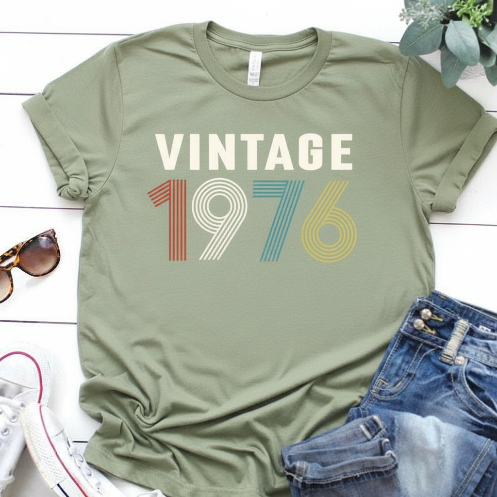 Green t-shirt with 'Vintage 1976' design on a white surface with jeans and sunglasses.