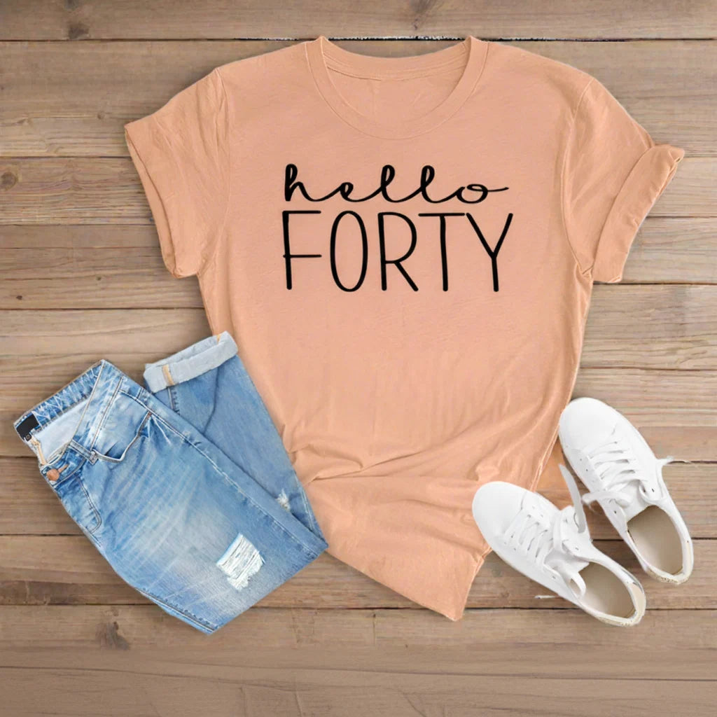 A peach-colored graphic t-shirt with 'hello FORTY' printed in black, styled in a flat lay with light blue denim shorts and white sneakers on a wooden floor background.