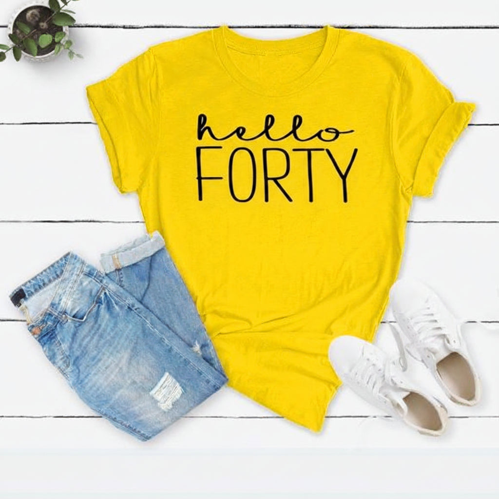 A bright yellow graphic t-shirt with the text 'hello FORTY' in black, styled in a flat lay with light blue jeans, white sneakers, and a small potted plant on a white wood plank background.
