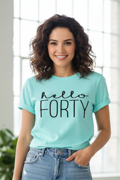 A woman with wavy brown hair smiling and wearing a light aqua graphic t-shirt that reads 'hello FORTY' in black script and block lettering, paired with blue jeans, set against a bright, softly blurred window background.