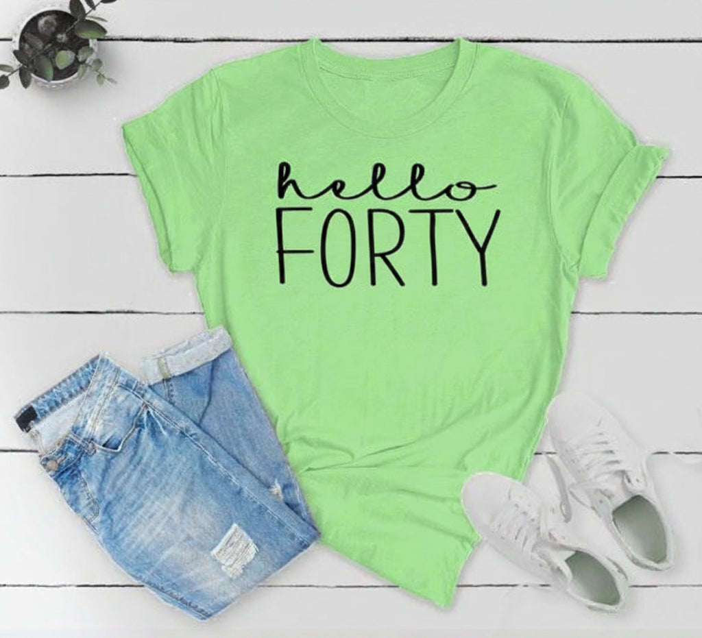 A bright lime green graphic t-shirt with 'hello FORTY' printed in black script and block lettering, styled in a flat lay with light-wash denim shorts, white sneakers, and a small potted plant on a white wood plank background.