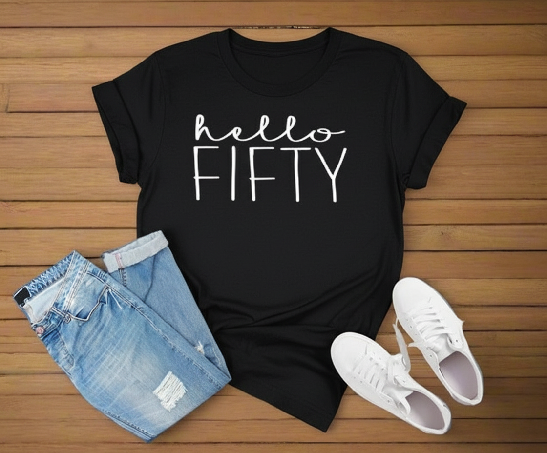 Black t-shirt with 'hello FIFTY' text, blue jeans, and white sneakers on a wooden surface