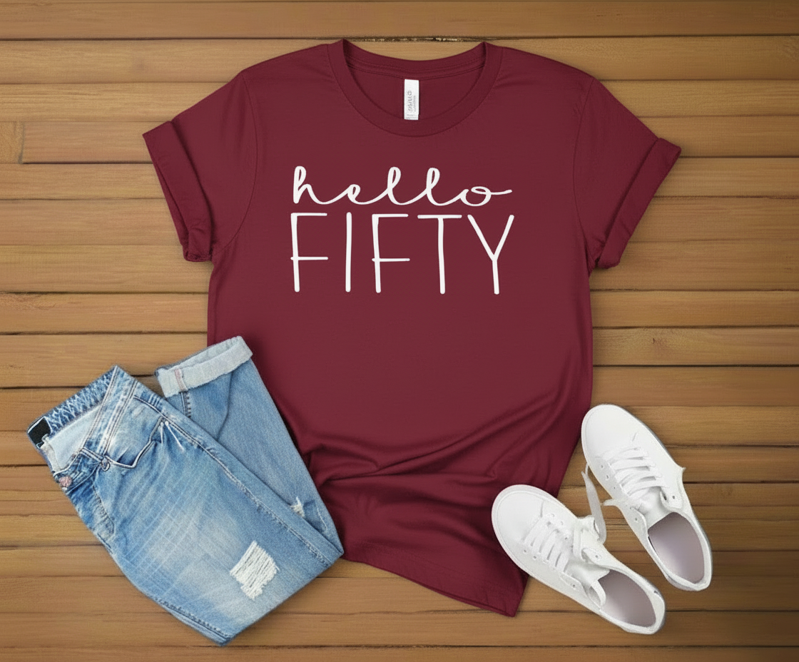 Maroon t-shirt with 'hello FIFTY' text, blue jeans, and white sneakers on a wooden surface